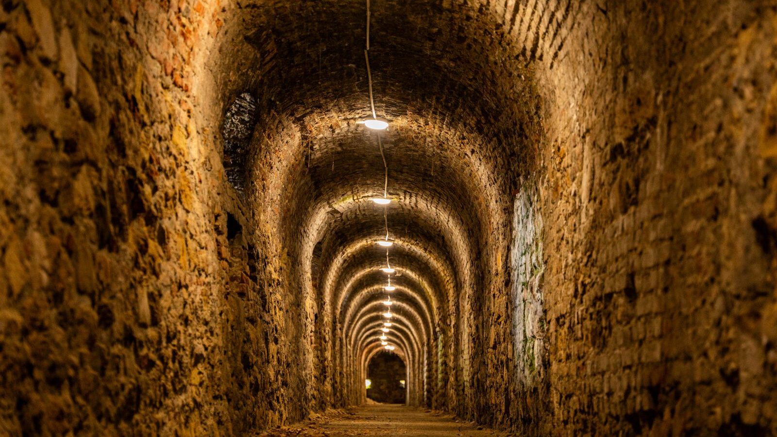A long, dimly lit ancient brick-vaulted underground passage receding into darkness, evoking the buried lower layers beneath the Basilica di San Clemente in Rome.