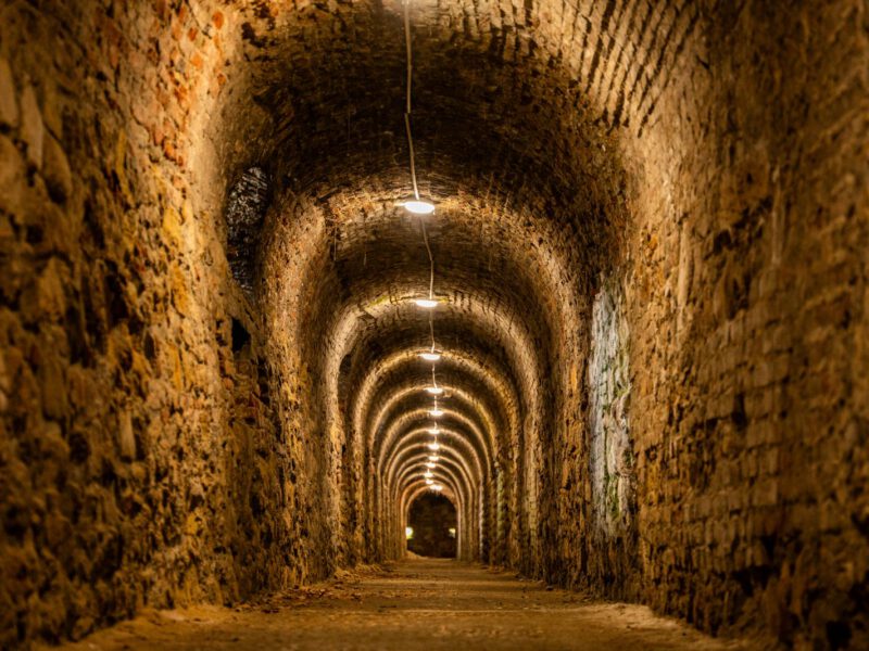 A long, dimly lit ancient brick-vaulted underground passage receding into darkness, evoking the buried lower layers beneath the Basilica di San Clemente in Rome.