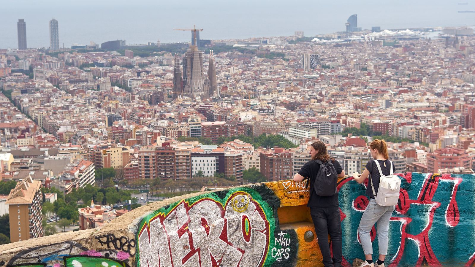 Panoramic view over Barcelona from the graffitied concrete platforms of the Bunkers del Carmel, with the Sagrada Família visible on the skyline.