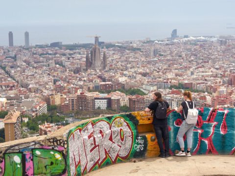 Panoramic view over Barcelona from the graffitied concrete platforms of the Bunkers del Carmel, with the Sagrada Família visible on the skyline.