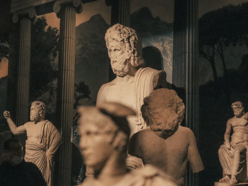 Ancient Roman marble statues of gods and figures dramatically lit against a dark museum backdrop, evoking the classical sculpture at Centrale Montemartini.