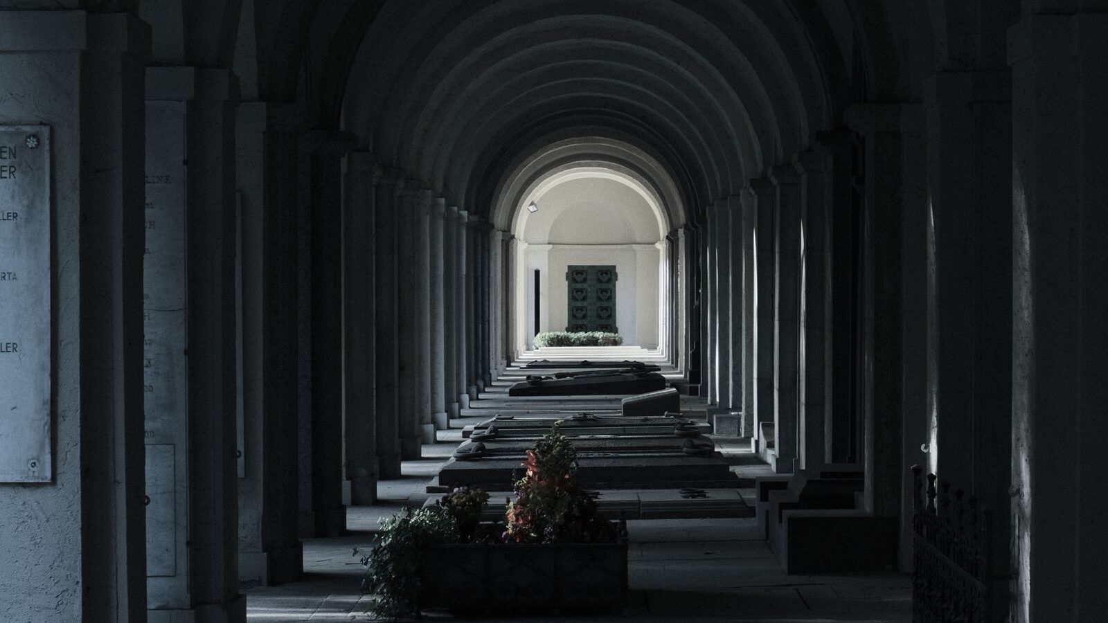 Dim vaulted stone arcade receding toward a distant lit archway, evoking a cool underground chamber.