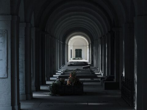 Dim vaulted stone arcade receding toward a distant lit archway, evoking a cool underground chamber.