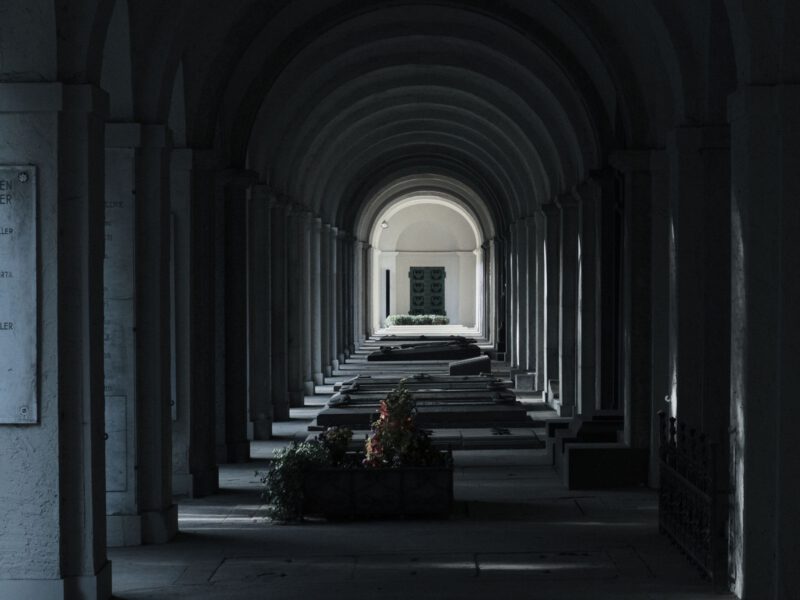 Dim vaulted stone arcade receding toward a distant lit archway, evoking a cool underground chamber.