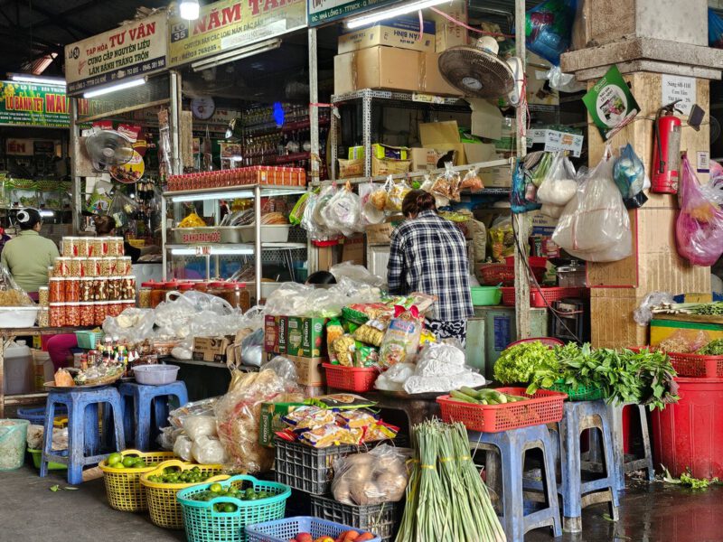 A busy Vietnamese market stall piled with fresh vegetables, herbs and goods, evoking the Dong Xuan Center's Asian wholesale halls in Berlin-Lichtenberg.