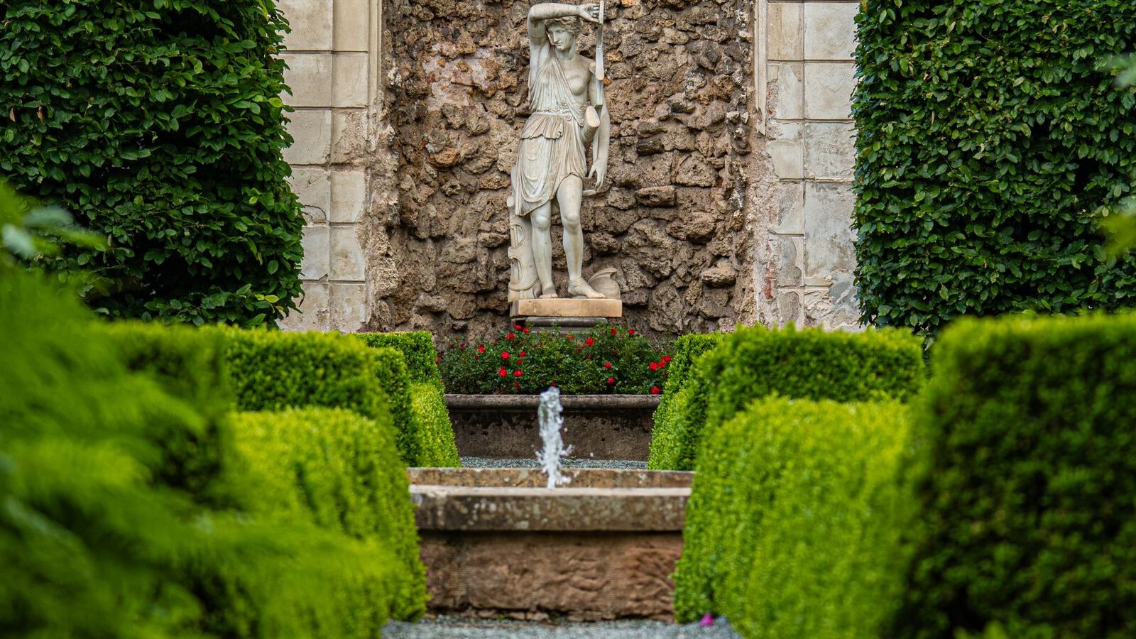 A classical stone statue in an arched garden niche with a small fountain, framed by clipped box hedges in a formal garden.