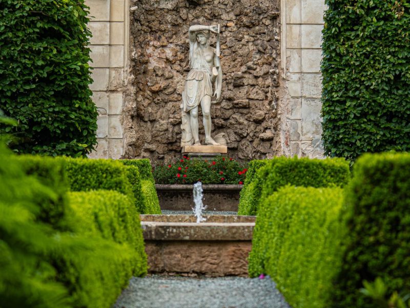 A classical stone statue in an arched garden niche with a small fountain, framed by clipped box hedges in a formal garden.