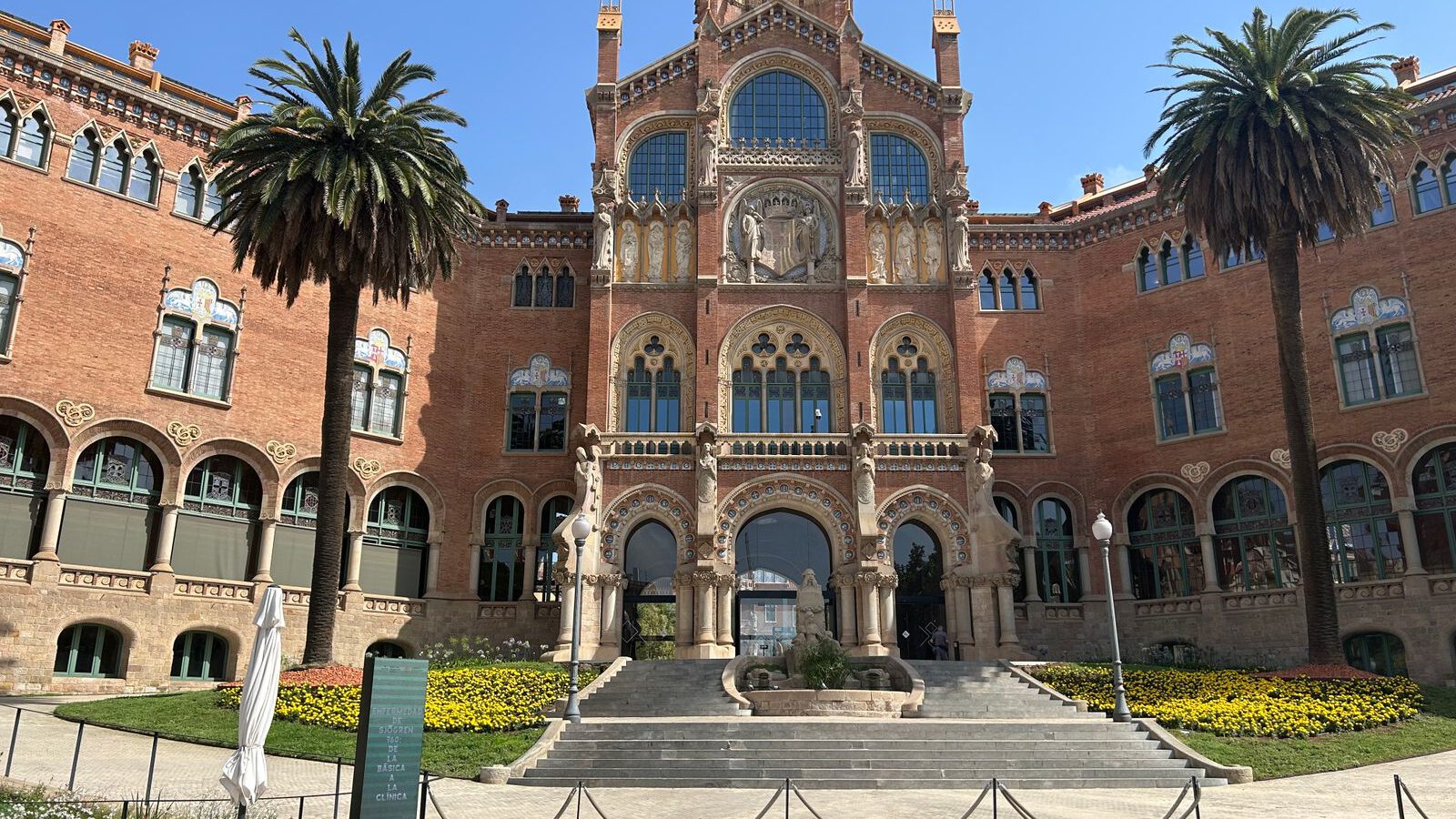 The ornate red-brick administration pavilion of the Recinte Modernista de Sant Pau in Barcelona, framed by palm trees under a blue sky.
