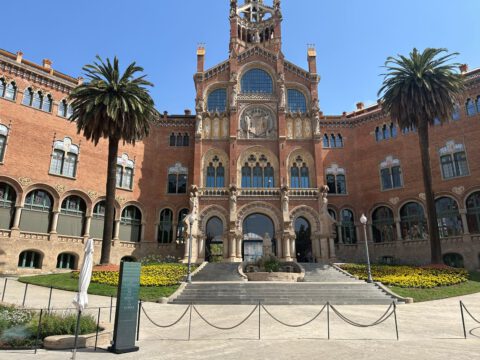 The ornate red-brick administration pavilion of the Recinte Modernista de Sant Pau in Barcelona, framed by palm trees under a blue sky.