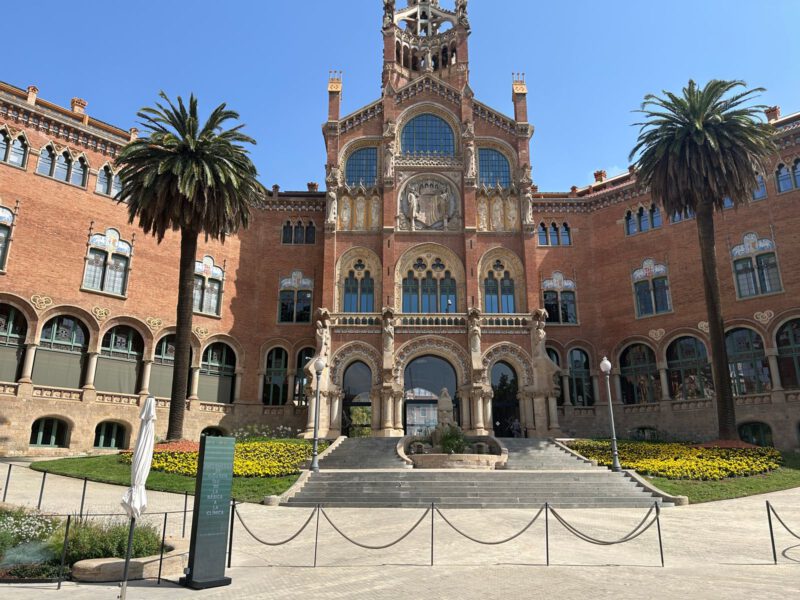 The ornate red-brick administration pavilion of the Recinte Modernista de Sant Pau in Barcelona, framed by palm trees under a blue sky.