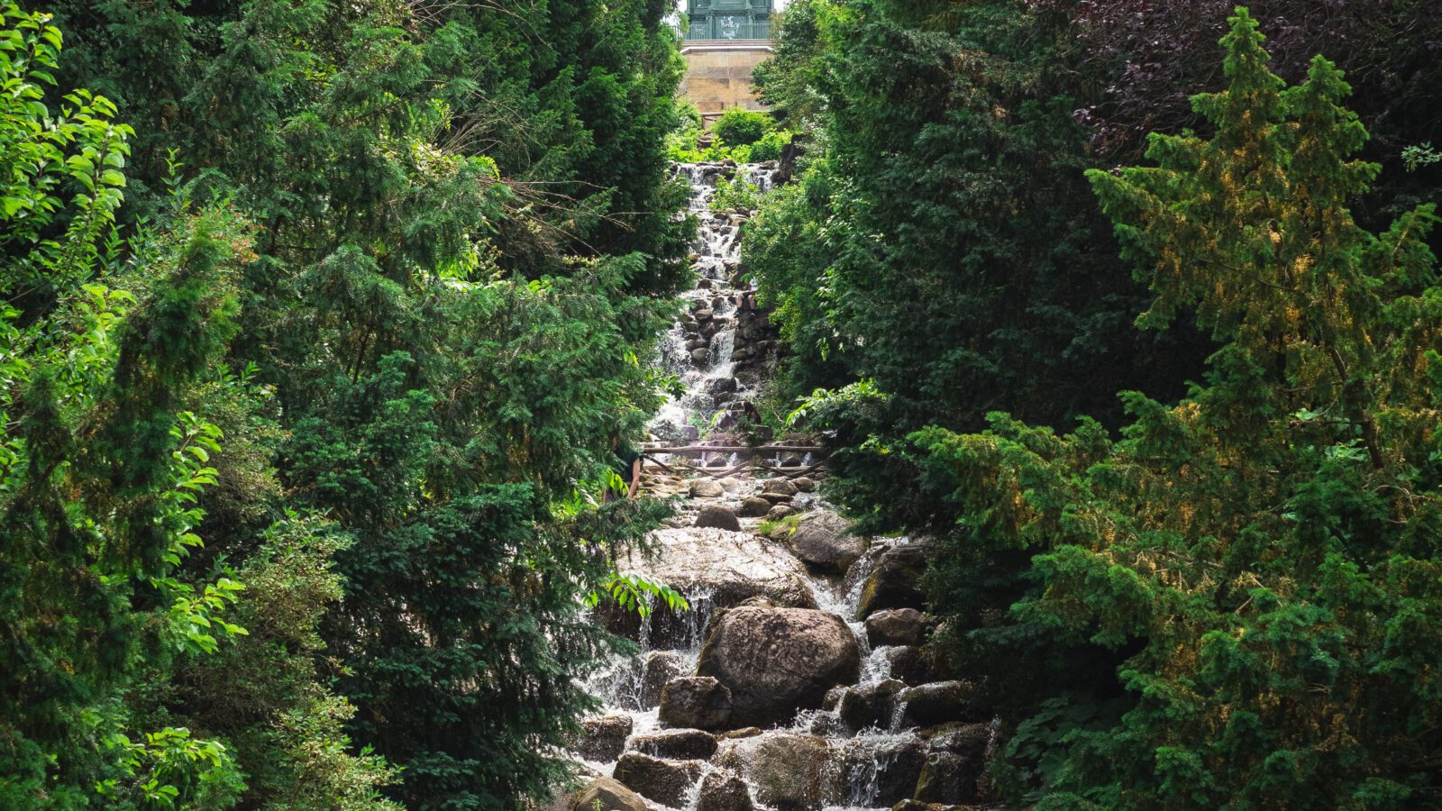 A tree-lined waterfall tumbling over rocks, evoking the Kreuzberg waterfall in Berlin's Viktoriapark.
