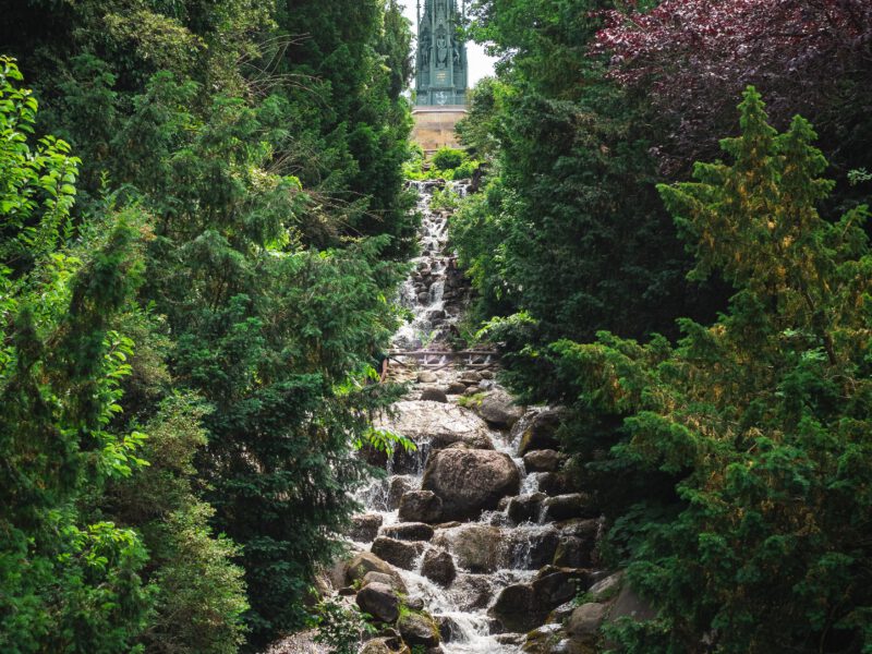 A tree-lined waterfall tumbling over rocks, evoking the Kreuzberg waterfall in Berlin's Viktoriapark.